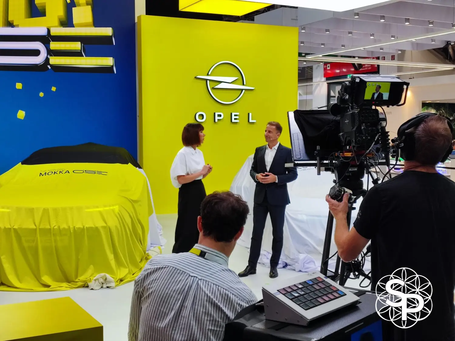 Photo montrant un tournage vidéo avec prompteur professionnel sur le stand Opel au Mondial de l'Auto. Au centre, une présentatrice et un homme en costume discutent. Derrière eux, une voiture neuve, The New Opel Mokka GSe, est recouverte d'un voile jaune et noir. Une grande caméra sur trépied les filme, gérée par deux membres de l'équipe technique au premier plan. Le fond est le logo Opel sur un mur jaune vif